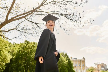 Neşeli üniversite mezunu cübbeli ve şapkalı güzel bir açık havada güneşli bir günün tadını çıkarıyor..