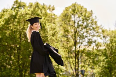 Gururlu genç bir mezun, ağaçlarla çevrili cübbesi ve şapkasıyla gülümsüyor..