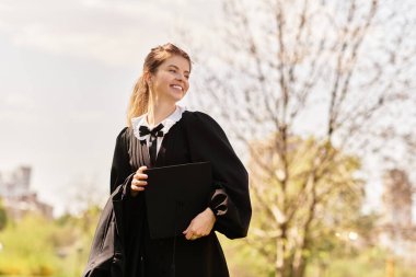 Genç bir üniversite mezunu yeşil bir parkta diplomasını tutarken sevinçle gülümsüyor..