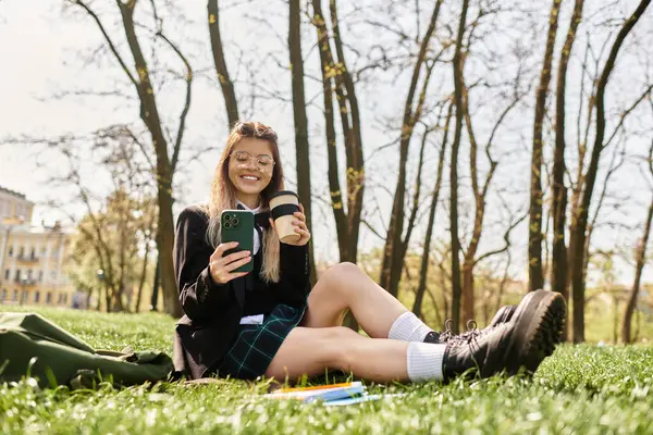 Genç ve güzel bir yetişkin, kahve ve telefonla çimlerin üzerinde dinleniyor, üniversitede güneşte ıslanıyor..