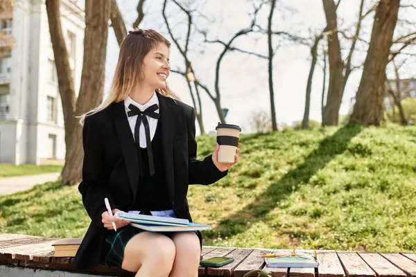 Genç kadın kitap ve kahveyle ahşap bankta oturuyor, güneşli bir üniversite ortamında gülümsüyor..