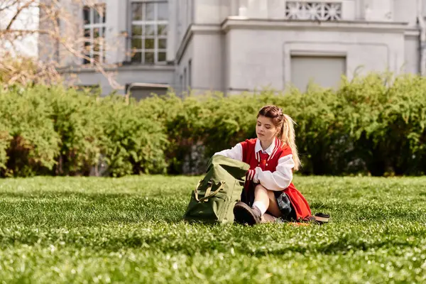 Genç bir öğrenci çimlerin üzerinde oturuyor, kırmızı bomba ceketini ve sırt çantasını ayarlıyor..