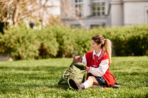 Young adult student sits on green grass, unpacking a bag while enjoying the warm sun.