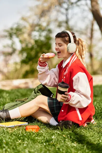 Bir üniversite öğrencisi çimenlerin üzerinde uzanır, tatlı bir ikram ve kahvenin tadını çıkarır..