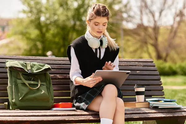Güzel bir üniversite öğrencisi, canlı bir park ortamında tabletine odaklanmış bir şekilde bankta oturuyor..