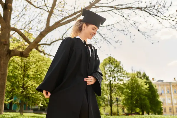 Mezuniyet elbisesi ve şapkasıyla dışarıda duran neşeli bir üniversiteli kız başarısını kucaklıyor..