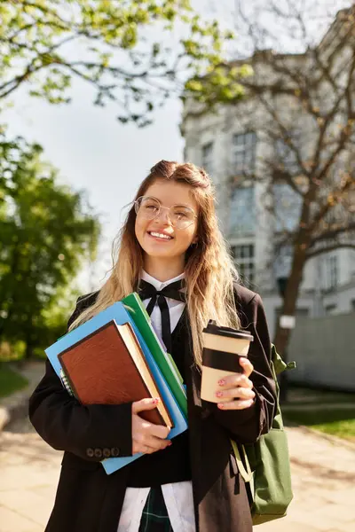 Şık üniformalı genç bir öğrenci ders kitabı ve kahvesini tutarken ışıl ışıl gülümsüyor..