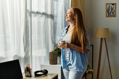 Genç bir kadın sıcak bir içecek tutarken mutlu bir şekilde gülümsüyor, evde pencereden dışarı bakıyor..