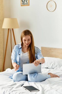 Neşeli genç bir kadın, mutlu bir şekilde yatağında oturuyor, tabletini parlak bir yatak odasında kullanıyor..