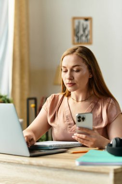 Genç bir kadın rahat bir ev ortamında görevlerine odaklanmak için bilgisayarını ve akıllı telefonunu kullanıyor..