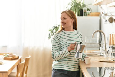 Genç bir kadın rahat bir şekilde giyinmiş, rahat mutfağında kahve yaparken gülümsüyor..