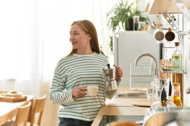 Güzel, genç bir kadın sıcak mutfağında duruyor, kahveyi yudumlarken mutlu bir şekilde gülümsüyor..