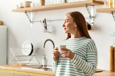 Evde sakin bir anın tadını çıkaran genç bir kadın, rahat mutfağında kahvesini yudumluyor, usulca gülümsüyor..