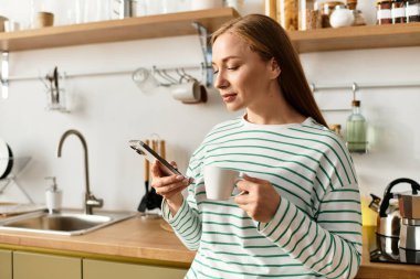Genç bir kadın telefonunu emerken fincanın tadını çıkarıyor ve evinde sıcak, rahat bir atmosfer yaratıyor..