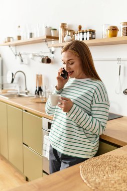 Huzurlu bir mutfak ortamında, genç bir kadın elinde sıcak bir fincan kahve tutarken telefonla konuşur..