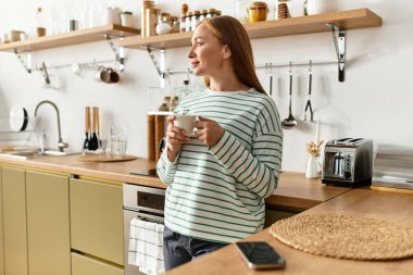 Güzel genç bir kadın rahat mutfağında duruyor, rahat bir gülümsemeyle ılık bir içkinin tadını çıkarıyor..