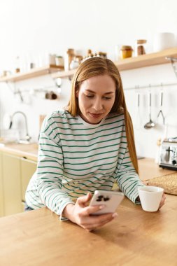 Genç bir kadın rahat mutfağında dinleniyor, akıllı telefonunu kontrol ederken çayını yudumluyor..