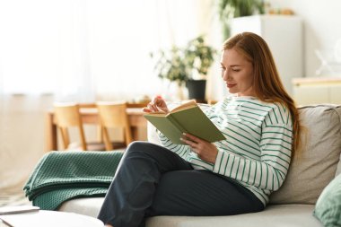 Genç bir kadın, rahat bir şekilde kanepede rahatça rahat rahat kitap okuyup rahat bir ev ortamında takılıyor..