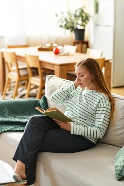 Genç bir kadın, doğal ışıkla çevrili iken rahat bir yaşam alanında okumaktan zevk alır..