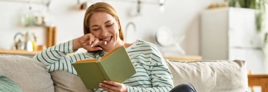 Genç bir kadın kanepede gülümsüyor, güzel bir kitaba dalmış ve rahat bir şekilde eğleniyor..