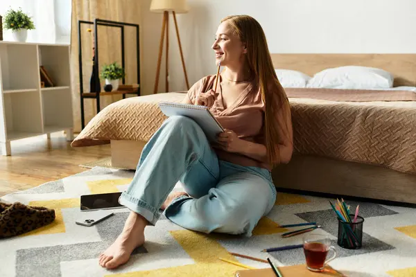 Genç bir kadın, rahat bir atmosferde renkli kalemlerle çevrili bir şekilde yerde çizimler yapıyor..