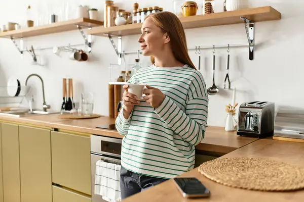 Güzel genç bir kadın rahat mutfağında duruyor, rahat bir gülümsemeyle ılık bir içkinin tadını çıkarıyor..