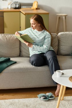 Genç bir kadın kanepede rahatlar, kitap okur ve sıcak bir evde sıcak bir içki yudumlar..