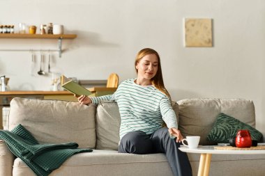 Genç bir kadın evinde rahat bir ortamda kitap ve sıcak bir içecekle rahatlamayı seviyor..