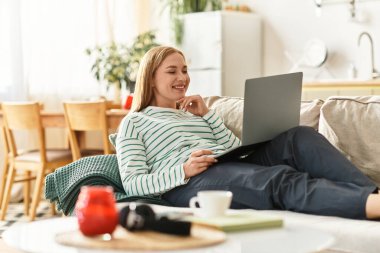 Genç bir kadın rahat bir kanepede dizüstü bilgisayarıyla rahat bir ortamda rahatlar..