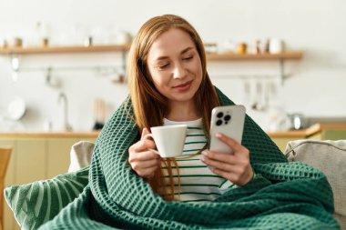 Sıcak bir battaniyeye sarılı genç bir kadın evde akıllı telefonunu kullanırken bir fincan kahvenin tadını çıkarıyor..