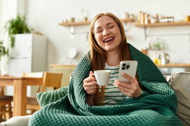 Genç ve güzel bir kadın bir kanepede rahatlar, elinde bardak tutarken gülümser ve telefonunu kullanır..