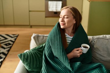 Sıcak bir yerde dinlenen genç bir kadın, yumuşak bir battaniyeye sarılı olarak kahvesini yudumluyor..