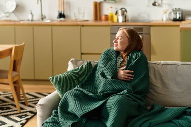 Genç bir kadın, evinde huzurlu bir öğleden sonra geçirir, rahat bir battaniyeye sarınır, nazikçe gülümser..