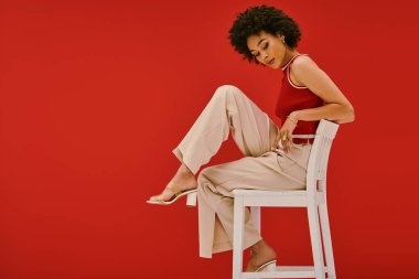 Büyüleyici genç bir kadın beyaz bir sandalyede otururken moda anlayışını sergiliyor..