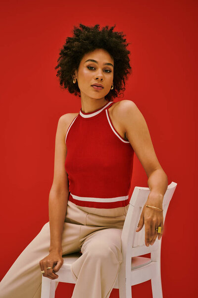 A confident young woman models fashion while seated against a vibrant red backdrop.