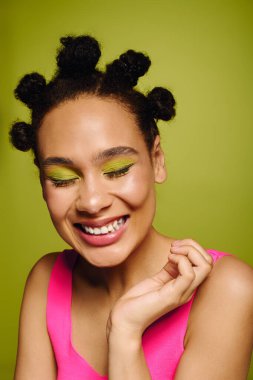 Genç bir kadın renkli makyajla neşeli bir poz veriyor ve parlak bir arkaplan karşısında eğlenceli bir saç stili..