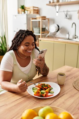 Genç bir Afrikalı Amerikalı kadın evde sağlıklı bir salatanın tadını çıkarıyor. Telefonunu kontrol ederken gülümsüyor..