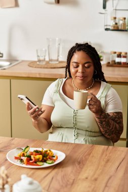 Genç bir Afrikalı Amerikalı kadın evde sıcak bir içecek yudumlarken taze salatanın tadını çıkarıyor..