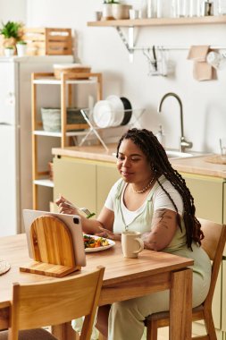 Genç bir Afrikalı Amerikalı kadın tabletini evde tutarken taze salata yiyor..