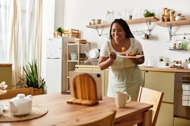 Gülümseyen genç kadın, mutfaktaki tabletle etkileşime girerken renkli bir salatanın tadını çıkarıyor..