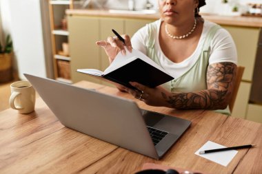 Odaklanmış genç bir kadın rahat ev ofisinde laptopuyla çalışırken notlar yazıyor..