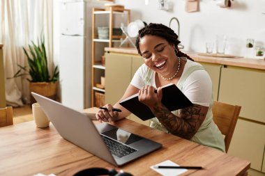 Genç siyah bir kadın mutfak masasında dizüstü bilgisayar ve not defteriyle çalışırken gülümsüyor..