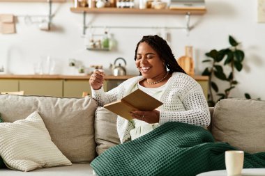 Genç bir kadın, evinde yumuşak bir battaniyeye sarılı olarak huzurlu bir sabah okumasının keyfini çıkarıyor..
