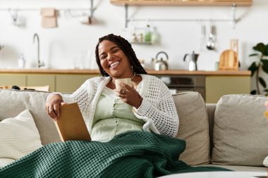 Genç bir Afrikalı Amerikalı kadın kanepesinde dinleniyor, çayını yudumluyor ve rahat evinde kitap okuyor..