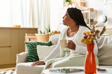 Genç bir Afrikalı Amerikalı kadın sıcak evinde pencereden dışarı bakarken çayını yudumluyor..