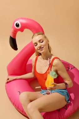 Genç bir kadın renkli bir tropikal içeceği yudumlarken pembe bir flamingo üzerinde dinleniyor..
