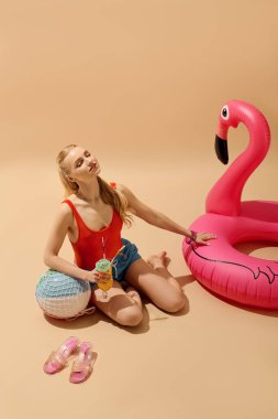 Canlı kırmızı bluzlu genç kadın yerde içki ve şakacı şişme flamingoyla rahatlıyor..
