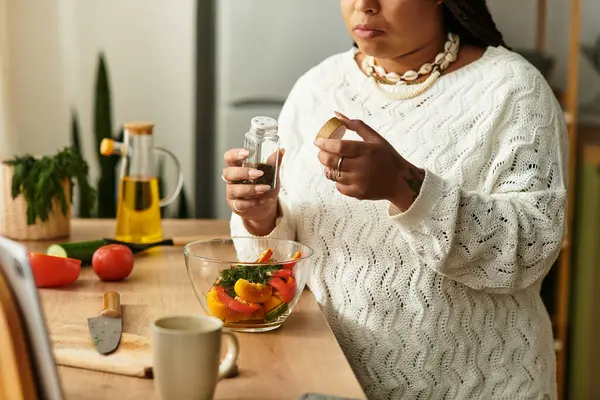 Genç bayan rahat mutfağında renkli bir salata hazırlıyor, lezzet katmak için baharat ekliyor..