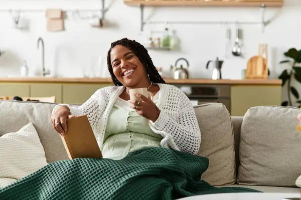 Genç bir Afrikalı Amerikalı kadın kanepesinde dinleniyor, çayını yudumluyor ve rahat evinde kitap okuyor..