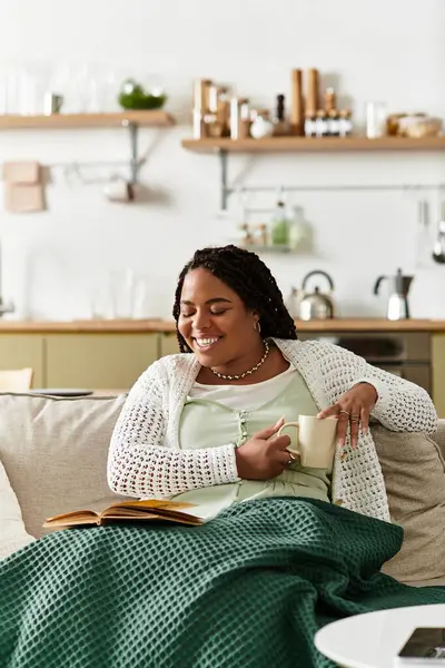 Genç bir kadın, çay içip kitap okurken battaniyeye sarılı sakin bir sabahın tadını çıkarıyor..